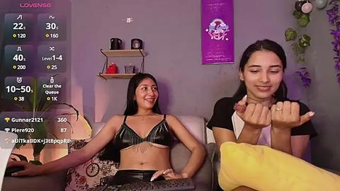 Media: Video of two women in a dimly lit room. One woman with long black hair in a black bra and shorts sits on a couch, smiling. The other woman, with long black hair, waves a yellow scarf, wearing a black shirt.