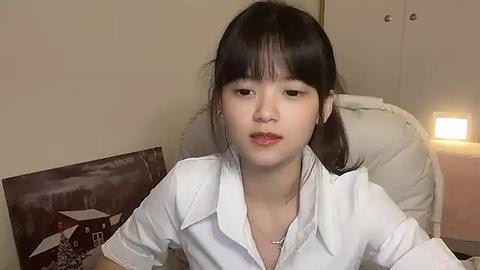 Media: Video of a young East Asian woman with straight black hair, wearing a white button-down shirt, sitting in a beige room with a wooden wall and a lit lamp.
