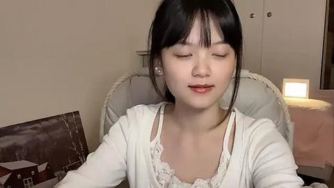 Media: Video of an Asian woman with fair skin, black hair, and bangs, wearing a white lace-trimmed top, sitting on a cushioned chair, eyes closed, serene expression. Background shows a beige wall and a framed picture.