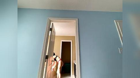 Media: Video of a child with light skin, dark hair, and wearing a pink shirt, bending over in a doorway, looking into a room with light blue walls, brown door, and white baseboards.