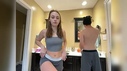 Media: Video of a young woman in a gray sports bra and shorts, standing confidently in a bathroom with beige walls and a mirror. Another person, shirtless, is reflected in the mirror.