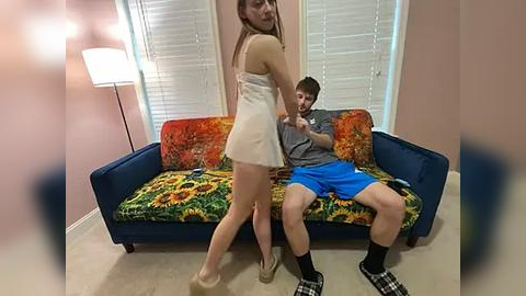 Media: Video: A young woman in a white dress and high heels stands behind a man in a gray shirt and blue shorts on a colorful couch.