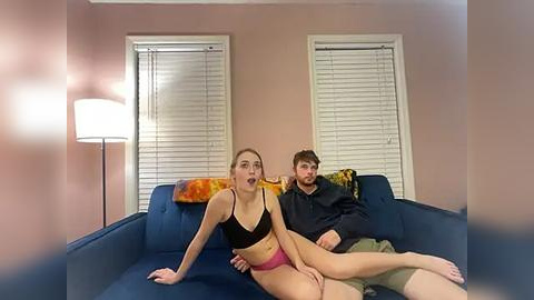 Media: Video of a young woman in a black sports bra and pink shorts, sitting on a blue couch, while a man in a black hoodie and glasses sits behind her, in a cozy, dimly lit room with beige walls and closed white blinds.