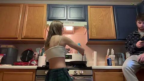 Media: Video of a woman in a white tank top and green shorts cooking in a small kitchen with wooden cabinets, a stove, and utensils on the counter. A man sits nearby in a black jacket, watching.