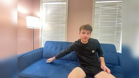 Media: Video of a young man with short brown hair, wearing a black long-sleeve shirt and shorts, sitting on a blue sofa, with a lamp and white blinds in the background.