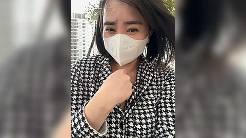 Media: Video of an Asian woman wearing a white mask, black houndstooth-patterned blouse, and holding her chin, blurred background of trees and buildings.