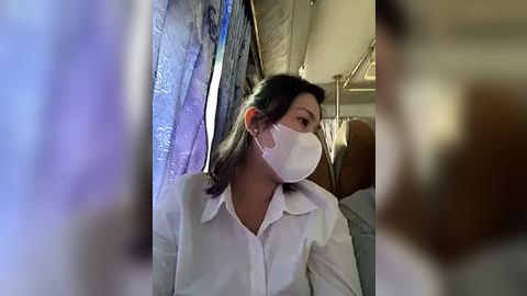 Media: Video of an Asian woman in a white surgical mask, white shirt, sitting in a hospital room with blue curtains and a metal bed frame.