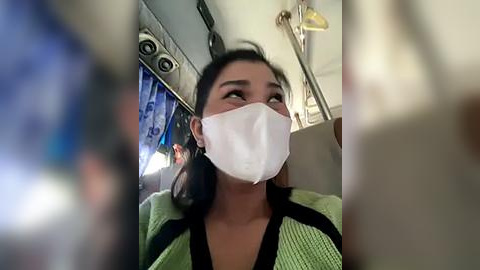 Media: Video of a woman in a green and black striped top wearing a white face mask, sitting on a bus with blurry background, overhead lights, and a TV screen.