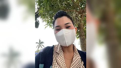 Media: Video of a woman with short black hair and light skin, wearing a white face mask, dark blue cardigan, and checkered shirt, standing outdoors under a palm tree.