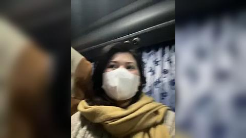 Media: A video of a woman wearing a yellow scarf, white face mask, and a beige sweater, sitting in a car with a blue patterned curtain in the background.