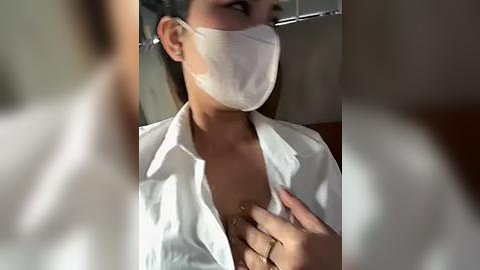 Media: Video of a woman with light skin, wearing a white surgical mask, white lab coat, and a necklace, looking down at her chest. The background is blurred, focusing on her attire and expression.