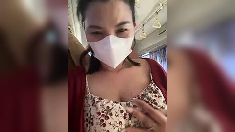 Media: Video of a woman wearing a white mask, red top, and floral patterned bra, inside a bus with blurred passengers in the background.