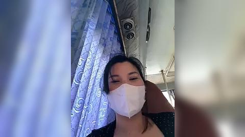 Media: A video of a woman with a mask, dark hair, and wearing a black top, sitting in a dimly lit, patterned room, possibly a hospital or clinic.