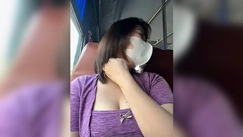 Media: Video of a woman wearing a face mask and purple top, adjusting her hair in a moving bus. Background shows blurred bus interior with metal railings.