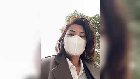 Media: Video of a woman with a white face mask, wearing a brown blazer over a white shirt, standing outdoors with blurred green foliage in the background.