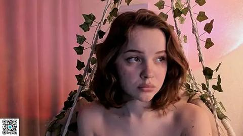 Media: Video of a fair-skinned, young woman with wavy brown hair, wearing a topless outfit, suspended in a green ivy swing, against a pink and white backdrop.