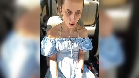 Media: Video of a young woman with light skin and brown hair, wearing a light blue off-shoulder dress, seated in a car.