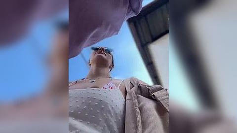 Media: A low-angle video of a woman with glasses, wearing a polka-dotted top and beige cardigan, standing under a blue sky.