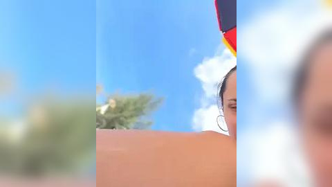 Media: Video of a woman with fair skin, dark hair, and hoop earrings, wearing a colorful hat with red, yellow, and blue stripes. She is partially visible, set against a bright blue sky with fluffy clouds.