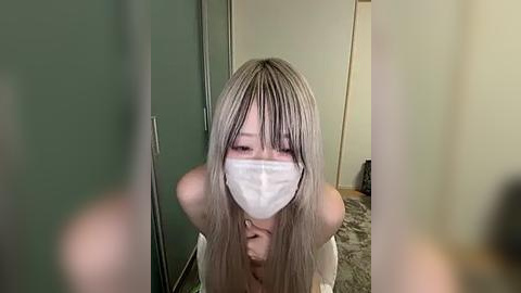 Media: Video of a woman with long, platinum blonde hair, wearing a white mask, standing in a dimly lit room with green walls, holding a phone.