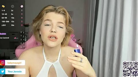 Media: Video of a blonde woman with light skin, wearing a white halter top, holding a blue lipstick. Background shows a pink gaming chair and a digital camera screen.