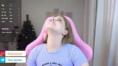 Media: A video of a young woman with light skin and short blonde hair, wearing a light blue t-shirt, reclining in a pink gaming chair, eyes closed, head tilted back. Background features a decorated Christmas tree.