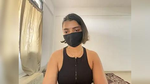 Media: A video of a young woman with medium brown skin, wearing a black face mask, black sleeveless top, and a beaded necklace, standing in a simple, brightly lit room with beige curtains and a patterned rug.