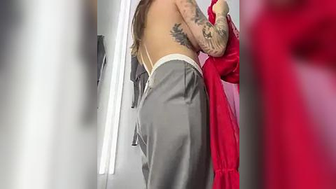 Media: Video of a topless woman with light skin, medium build, and tattoos, wearing loose gray sweatpants and holding a red garment, standing in a brightly lit room with gray walls.