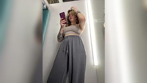 Media: Video of a plus-sized, tattooed, Caucasian woman with blonde hair in a beige top and high-waisted gray pants taking a selfie in a narrow, brightly-lit dressing room.