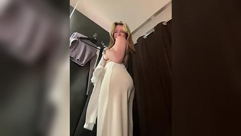 Media: Video of a blonde woman in a white dress, standing in a dimly lit dressing room with dark curtains, holding a towel over her face.