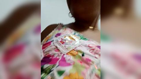 Media: Video of a woman with dark skin wearing a colorful, floral-patterned dress, with a gold necklace and a small, shiny object attached to her chest.