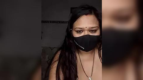 Media: A video of a South Asian woman with long black hair, wearing a black face mask, gold necklace, and a black top, set against a dimly lit bathroom background.