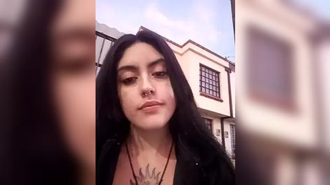 Media: Video of a young woman with long black hair, fair skin, and a nose piercing, wearing a black jacket. Background features a suburban house with a window and partly cloudy sky.