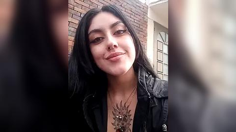 Media: Video of a young woman with long black hair, fair skin, and a nose ring, smiling against a brick wall. She wears a black jacket and a floral tattoo on her chest.