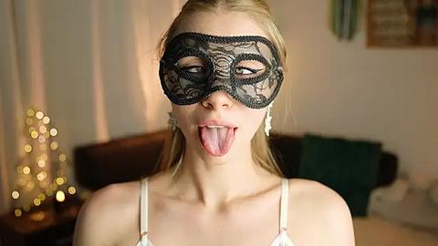 Media: Video of a young woman with fair skin, blonde hair, wearing a black lace masquerade mask, sticking her tongue out playfully. She's in a cozy, dimly lit room with a Christmas tree and green sofa in the background.