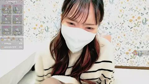 Media: Video of an Asian woman with long, straight dark hair wearing a white face mask, striped sweater, and lying on a white surface, with floral wallpaper in the background.