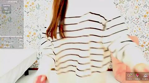 Media: Video of a woman with long, straight red hair, wearing a white and black striped sweater, sitting on a bed with white sheets. The background features floral-patterned wallpaper.