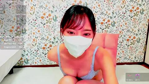 Media: A video of an Asian woman with short black hair, wearing a light blue lace bra, and a white face mask, sitting on a chair with a floral wallpaper background, taken from a live streaming perspective.