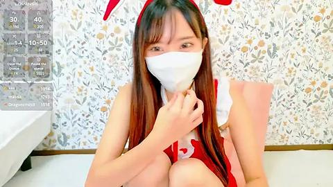 Media: Video of a young Asian woman with long brown hair, wearing a red bunny outfit, white face mask, and floral wallpaper background.