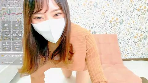 Media: Video of an Asian woman with long, straight brown hair, wearing a white face mask and a light brown sweater, kneeling on a white couch against a floral wallpaper background.