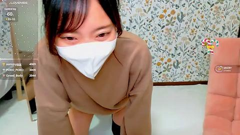 Media: A video of an Asian woman in a beige coat, wearing a white mask, bent over, in a room with floral wallpaper and a pink chair.