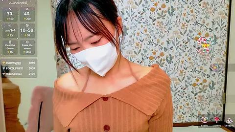 Media: Video of an East Asian woman with medium skin tone, wearing a beige button-up blouse and white mask, standing in a room with floral wallpaper.