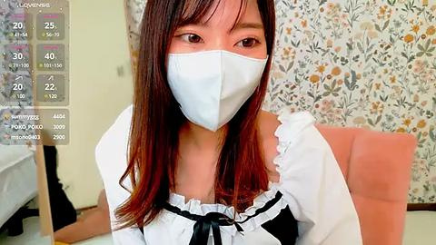 Media: Video of a young Asian woman in a white frilled dress with a black bow, wearing a face mask, sitting indoors with floral wallpaper.