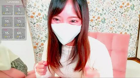 Media: Video of an Asian woman with long, straight black hair, wearing a white face mask, white sweater, and a pink blanket in a floral-patterned room.