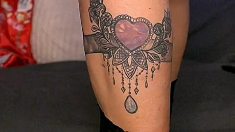 Media: A video showcasing a detailed, black and white tattoo on a light-skinned forearm featuring a heart with a gemstone center, surrounded by intricate mandala patterns and hanging teardrop shapes. The background is blurred, revealing a red and white floral pattern.