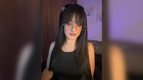 Media: Video of a young woman with long black hair, wearing glasses, a black sleeveless top, and a necklace, indoors.