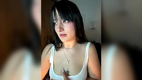 Media: Video of a young woman with long black hair, wearing a white tank top, sitting indoors. She has tattoos on her chest and neck, and appears to be in a thoughtful, introspective pose.