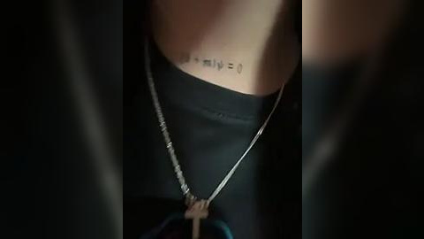 Media: A close-up video of a person wearing a black t-shirt with a small tattoo on their neck. The person is also adorned with a silver chain necklace. The background is dark, with minimal lighting, focusing attention on the tattoo and necklace.