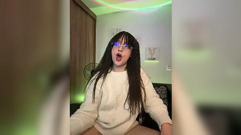 Media: A video of a young woman with long black hair, wearing a white knitted sweater and glasses, sitting in a dimly lit room with green and red LED lights.