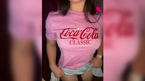 Media: Video of a woman with long brown hair, wearing a pink T-shirt with \"Coca-Cola Classic\" in red cursive, lifting her skirt, revealing her thighs.